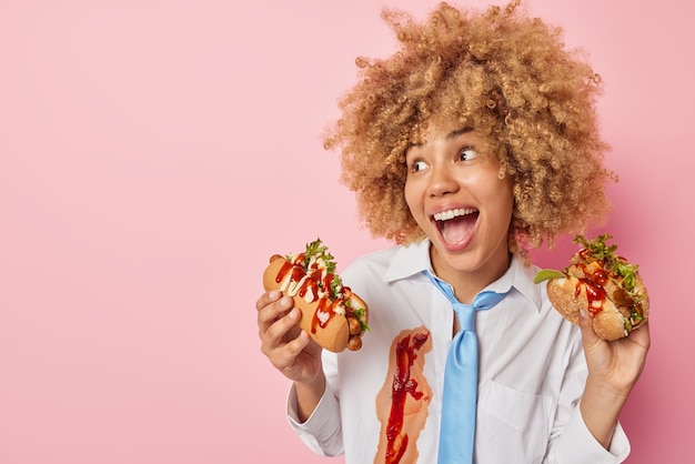 Cheerful woman exclaims loudly looks happily eats fast food\
holds hot dog and burger wears formal shirt and tie isolated over\
pink background blank space for your advertising content binge\
eating