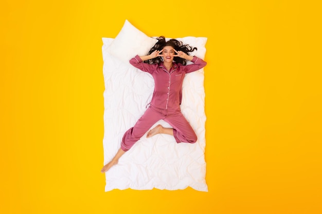 Cheerful woman gesturing peace signs lying on blanket yellow background