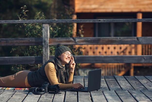 Cheerful woman in glasses lies on wooden terrace and works on laptop concept of freelance remote work on Internet