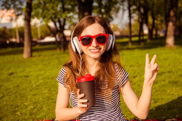 Cheerful woman in glasses listening music and gesturing while drinking coffee in the park