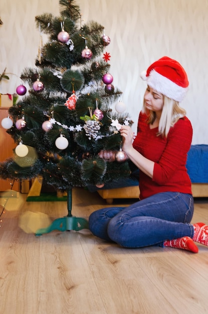 아늑한 집에서 크리스마스 트리 근처에서 즐거운 시간을 보내는 쾌활한 여자. 머리에 빨간 스웨터와 사슴 뿔
