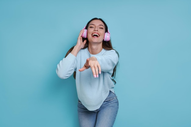 Cheerful woman in headphones pointing camera while standing against blue background