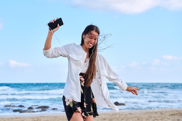 Cheerful woman in headphones with smartphone listening to music dancing on beach