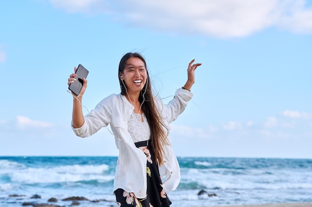 Cheerful woman in headphones with smartphone listening to music dancing on beach