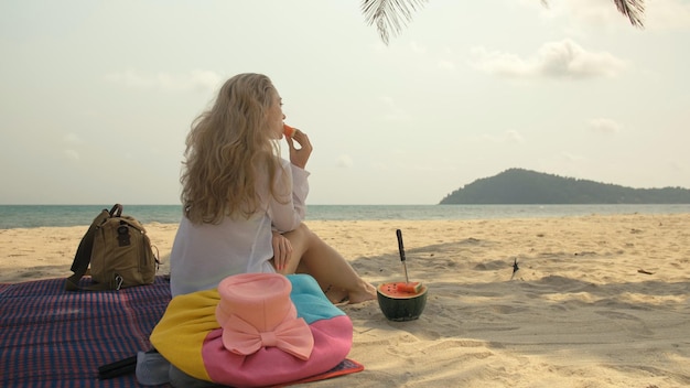 熱帯の砂浜の海でスイカのスライスを保持し、食べる陽気な女性