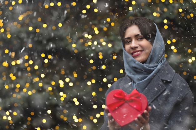 Cheerful woman holding red gift box in a heart shape during the snowfall. Empty space