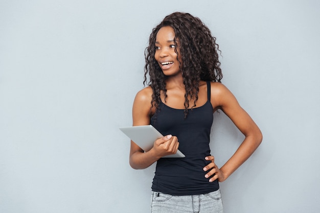 Cheerful woman holding tablet computer and looking away over gray wall