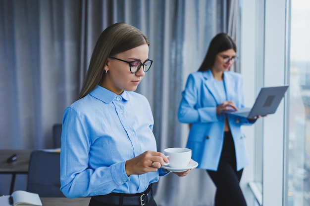 Веселая женщина в классических очках улыбается в свободное время в офисе с кофе позитивная европейская женщина в синей рубашке стол с ноутбуком удаленная работа коллега на заднем плане избирательный фокус