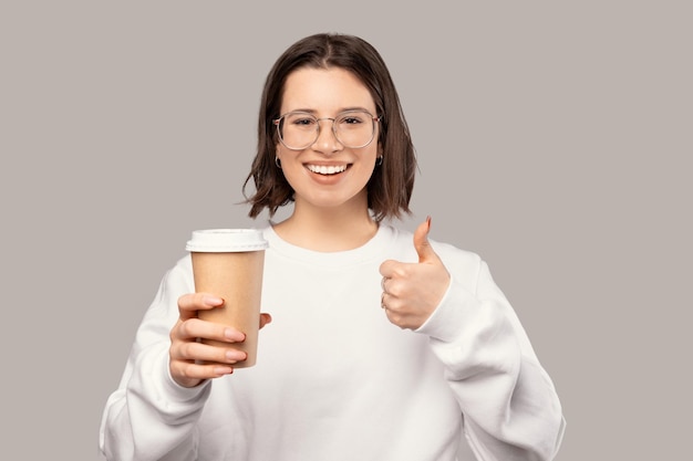 Photo cheerful woman is holding a disposable paper cup while showing thumb up