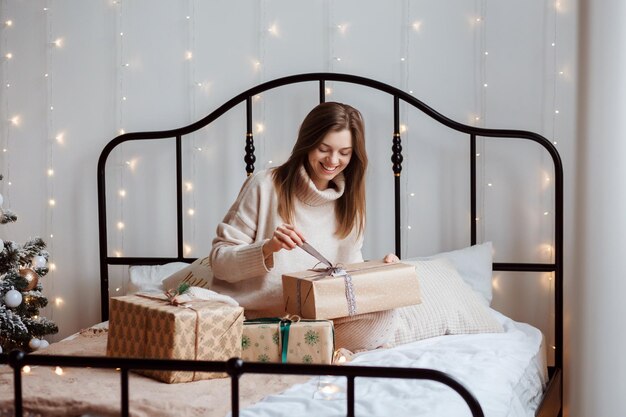 Cheerful woman is unwrapping Christmas or New Year gifts sitting on the bed in the festively decorated room.