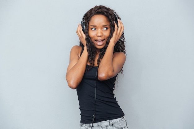 Cheerful woman listening to music in headphones and looking away over gray wall