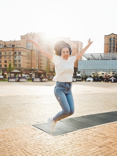 A cheerful woman listens to music through headphones outside on a sunny day a girl with headphones jumps on the street generation z