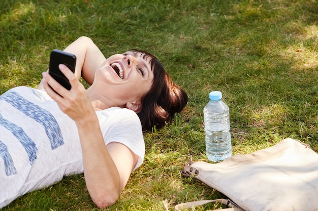 Donna allegra che si trova nell'erba nel parco con il telefono