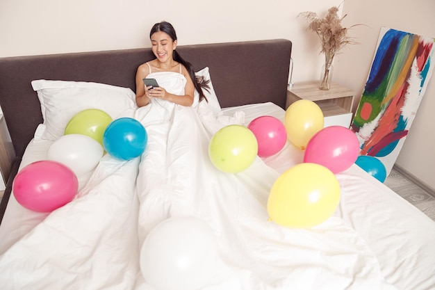 Веселая женщина лежит в постели с воздушными шарами и пользуется мобильным телефоном