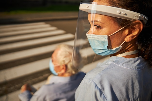 Cheerful woman medicine worker in blue uniform walking with mature disable man in the outdoors