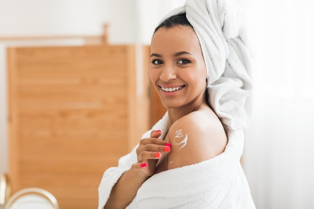 Cheerful woman moisturizing body applying moisturizer on shoulders in bathroom