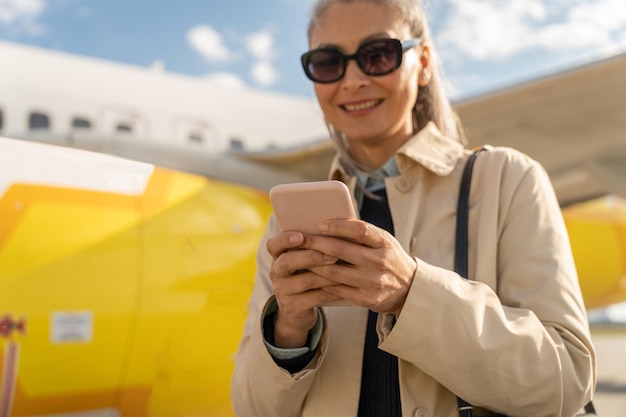 飛行機の近くの空港で屋外に立っている電話を使用してサングラスをかけた陽気な女性の乗客