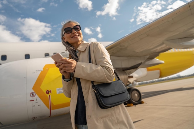 Cheerful woman passenger in sunglasses using phone standing outdoors at airport near plane