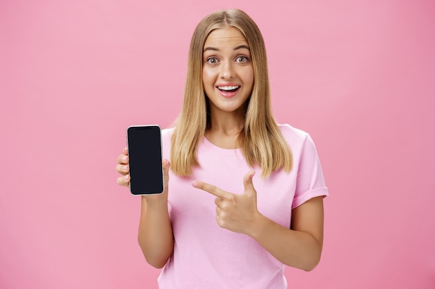 ピンクの背景の上に立って面白がって感動した笑顔のデバイス画面を指している陽気な女性。
