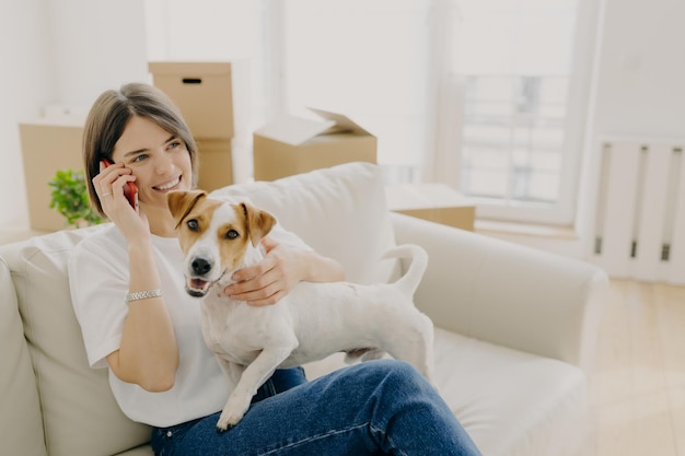 Cheerful woman poses on comfortable sofa speaks with friend via smartphone plays with pet shares impression and good news about buying new flat cardboard boxes in background Delivery service