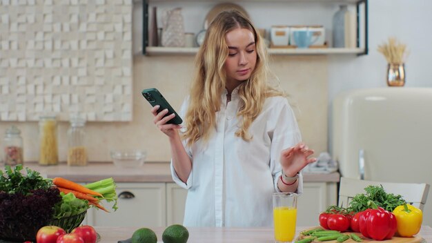 野菜の食事を準備する陽気な女性、スマートフォンでオンラインレシピを検索してチェックする ヘルスケアのコンセプト オーガニックの新鮮な果物