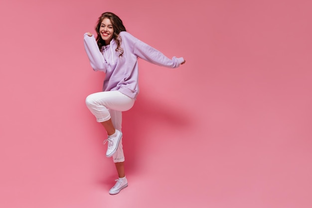 Cheerful woman in purple oversized hoodie and white pants moves on isolated Charming brunette girl smiles and dances on pink background