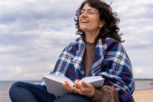 A cheerful woman reads a book on the seashore covered with a blanket