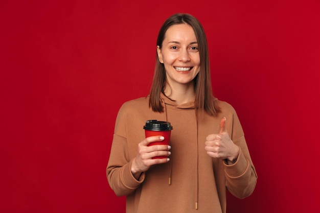 Cheerful woman recommends this coffee white holding a to go cup and thumb up