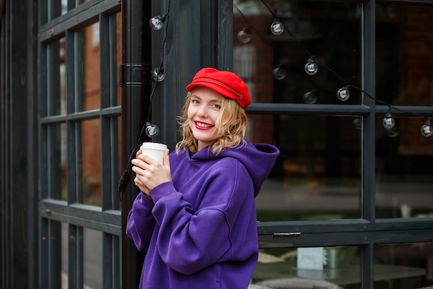 Cheerful woman in red cap and violet hoodie is drinking coffee outdoors, coffee break.