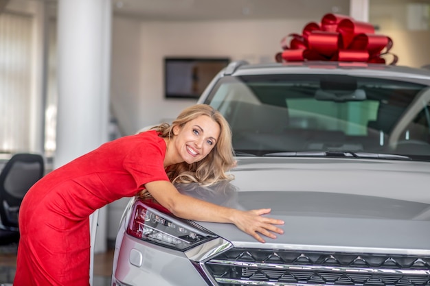 Cheerful woman in red dress hugging new car