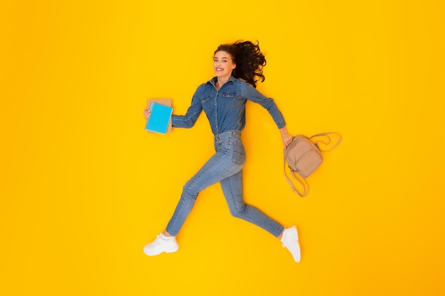 Cheerful woman running holding backpack and copybooks over yellow background