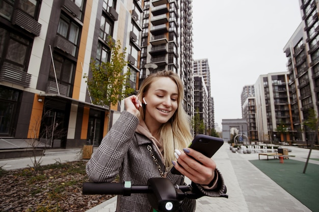 Cheerful woman on scooter wearing casual cozy coat, fixing her hair and chatting by her smartphone with electric transport rental service. Apartment blocks on background. Good service concept.