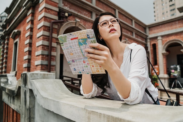 Cheerful woman searching direction on location paper map while
traveling abroad in summer. happy female tourist visit foreign
city. young girl leaning on railing of traditional chinese
building