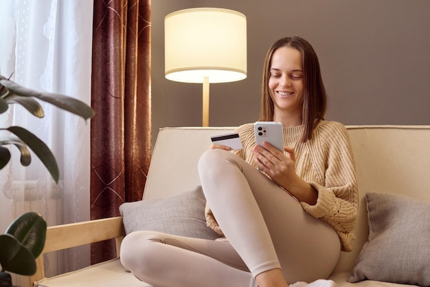 Cheerful woman shopper purchasing goods or services in internet store holding smartphone and banking credit card involved in online mobile shopping at home