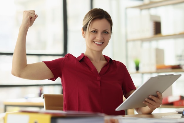 Cheerful woman shows arm muscles holding clipboard