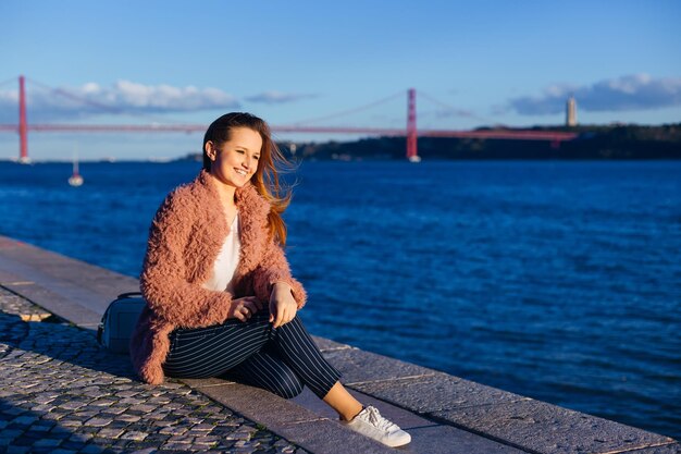 Cheerful woman sits on the river bank in sunny weather in spring The girl is dressed in a pink coat