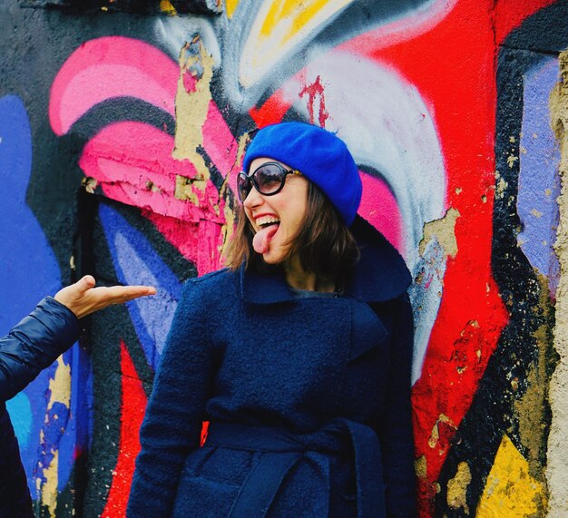 Photo cheerful woman sticking out tongue against colorful graffiti wall