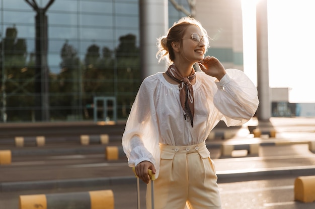 Cheerful woman in stylish blouse and beige pants smiles widely Attractive blonde girl in white shirt poses with luggage near airport