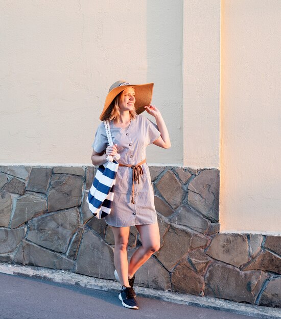 Cheerful woman in summer clothes walking on the street holding her hat in sunset light