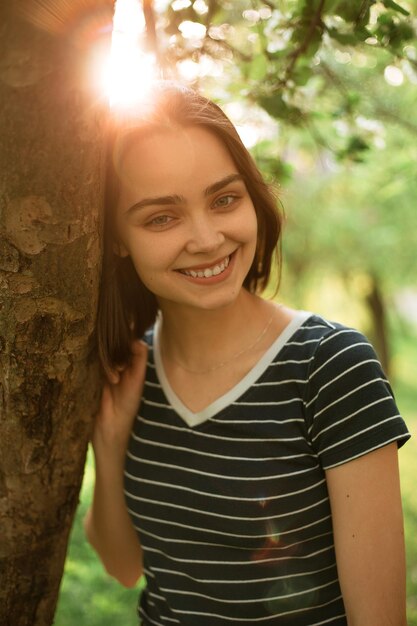 Cheerful woman in summer outfit standing near tree in park and looking at camera