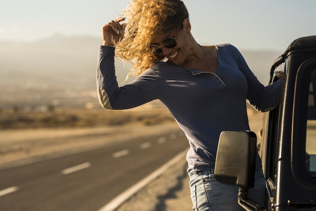 Cheerful woman in sunglasses hanging on side door of car at roadside. Young woman enjoying vacation on road trip. Carefree woman having fun leaning on outside of car door on her vacation trip