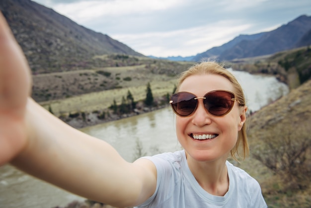 Cheerful woman in sunglasses takes a selfie on background of mountain landscape. Active lifestyle.