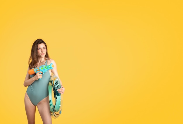 Cheerful woman in swim clothes posing with water gun and inflatable ring having fun on yellow