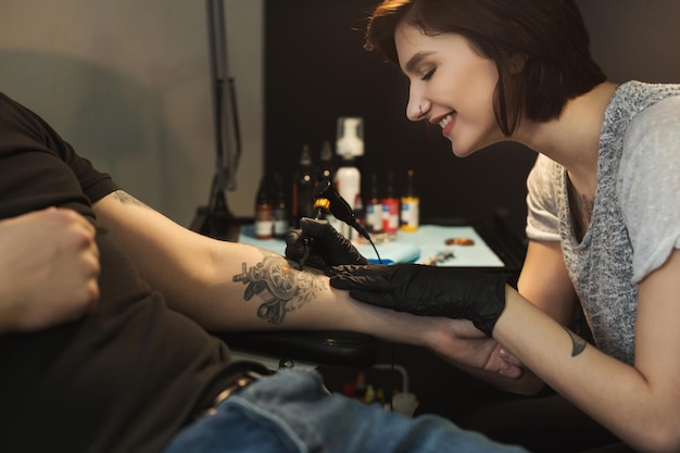 Cheerful woman tattooist doing tattoo with tattoo machine on male arm. Unusual female profession, copy space