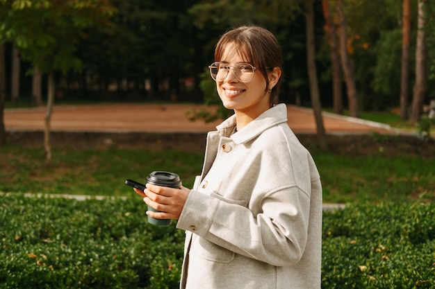 공원에서 산책하는 쾌활한 여성은 전화기와 뜨거운 커피 한 잔을 들고 있습니다.