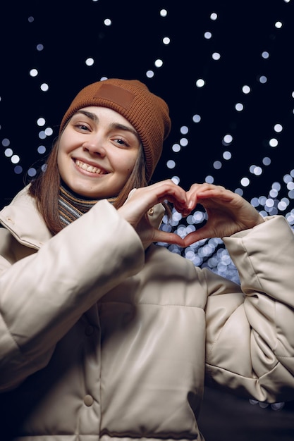 Cheerful woman in warm winter clothes showing heart gesture or love sign while looking at camera