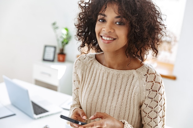 Cheerful woman wearing in casual clothes holding smartphone and looking at the camera while standing near the table at office