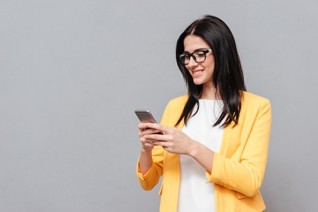 Cheerful woman wearing eyeglasses and dressed in yellow jacket chatting by her phone over grey surface. Look at phone.