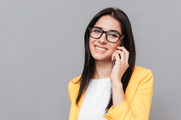 Cheerful woman wearing eyeglasses and dressed in yellow jacket talking by her phone over grey surface. Look at front.