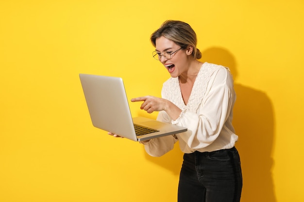 Cheerful woman wearing eyeglasses is using laptop computer on yellow background